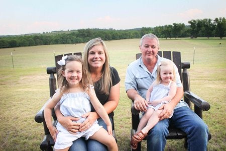 Travis and Kelly Cushman sit in adirondack chairs holding their young daughters.