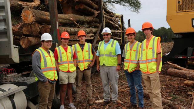 Group of (mostly young) men in yellow vests and hard hats on timber land